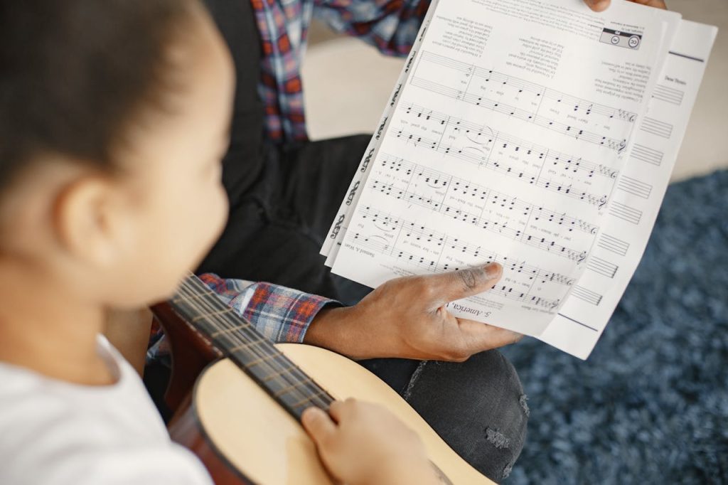 A young girl learning guitar with a teacher, focusing on a music sheet.
